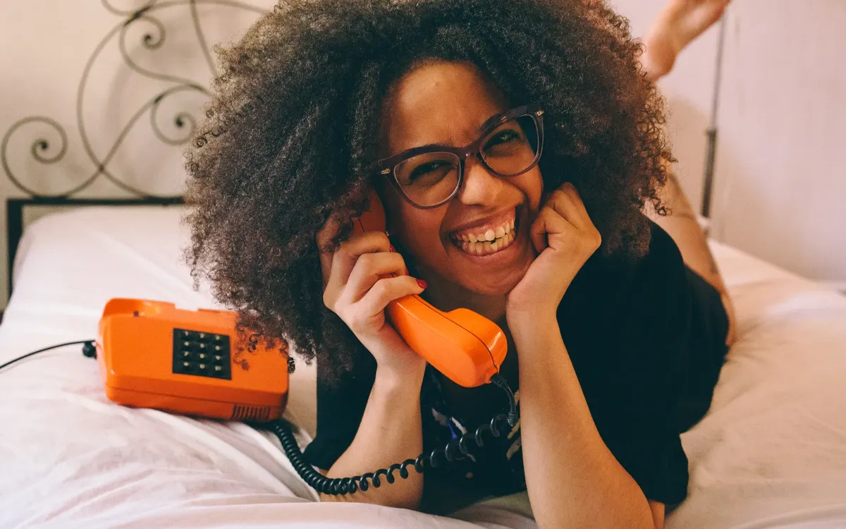 Cheerful woman lying on bed, holding a vintage orange phone, smiling joyfully.